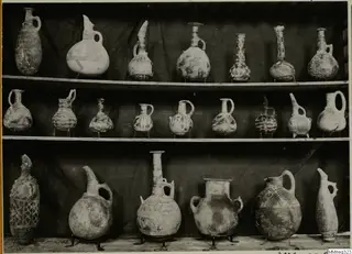 Three shelves of painted pottery jugs and juglets against a dark draped cloth