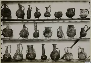 Three wooden shelves holding about twenty pottery jugs and two-handled jars, most standing on thin wire supports.