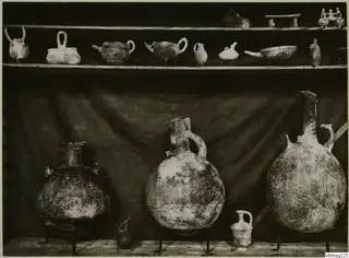 Pottery vessels on two shelves and metal stands in front of a draped dark cloth.