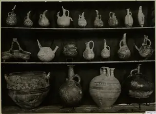 Painted pottery vessels, mostly jugs and juglets, arranged on three wooden shelves in front of a dark cloth.
