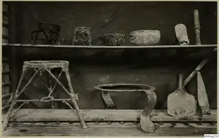 Corroded metal frames, bowls, a flask and a blade on two wooden shelves against a dark cloth.