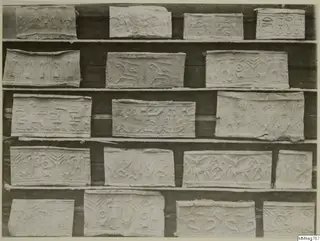 Rows of about eighteen pale rectangular pieces with shallow relief designs, set on dark shelves