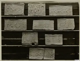 Ten pale rectangular impressions with stamped designs of figures and signs, arranged on wooden shelves