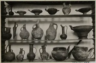 Three wooden shelves holding many pottery jugs, jars and footed bowls, displayed in the Cyprus Museum
