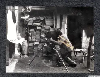 Man in a suit photographing with a large camera on a tripod, with stacked crates and boxes behind him