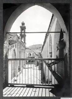 Cobbled courtyard with a church bell tower and a staircase, seen through an arched opening with iron railings