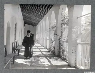 Bearded man in a long black robe and black cap standing on a paved walkway beside a row of white arches