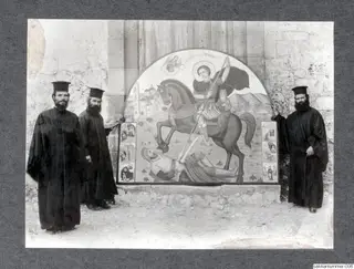 Three bearded men in black robes and tall black hats stand before a painted arched icon of a haloed rider spearing a fallen armoured knight.