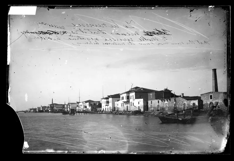 Row of stone and plastered houses along the Larnaca waterfront, with a boat moored in the foreground and a tall chimney at right.