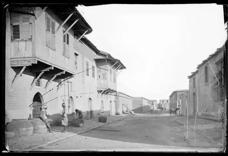 Unpaved street in Larnaca lined with two-storey houses, shuttered balconies, large clay jars and a few men
