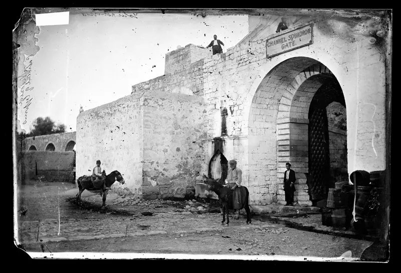 Stone gateway with a tall arch and a sign above it, with two donkeys and riders in a cobbled street
