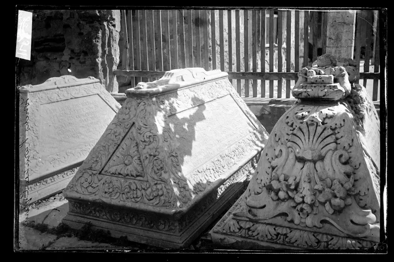 Three carved stone tombs with foliage decoration and inscribed panels, in front of a wooden fence