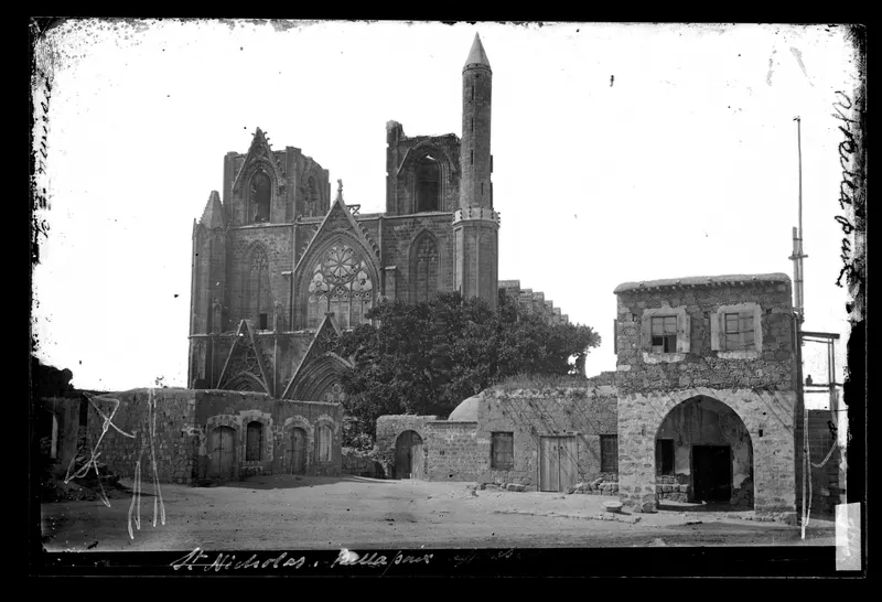 West front of the cathedral of St Nicholas in Famagusta, with a tall minaret, beside a dirt street and low stone houses.