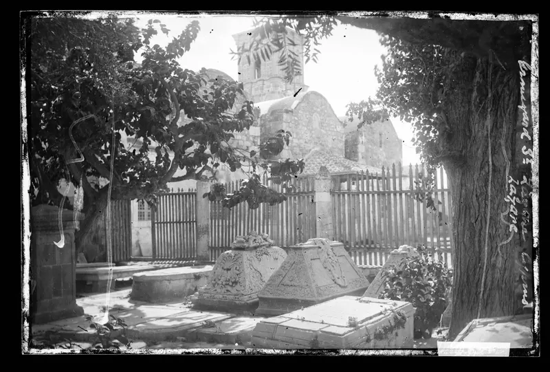 Carved stone tombs in a graveyard under trees, with a church with a tower behind a wooden fence