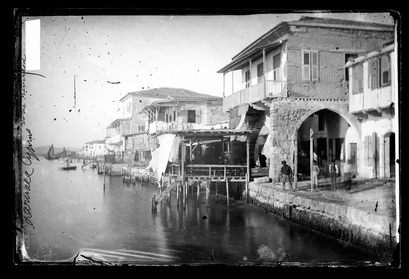 Two-storey stone houses with wooden balconies and shutters stand over the water at Larnaca, with figures on the quay at right.