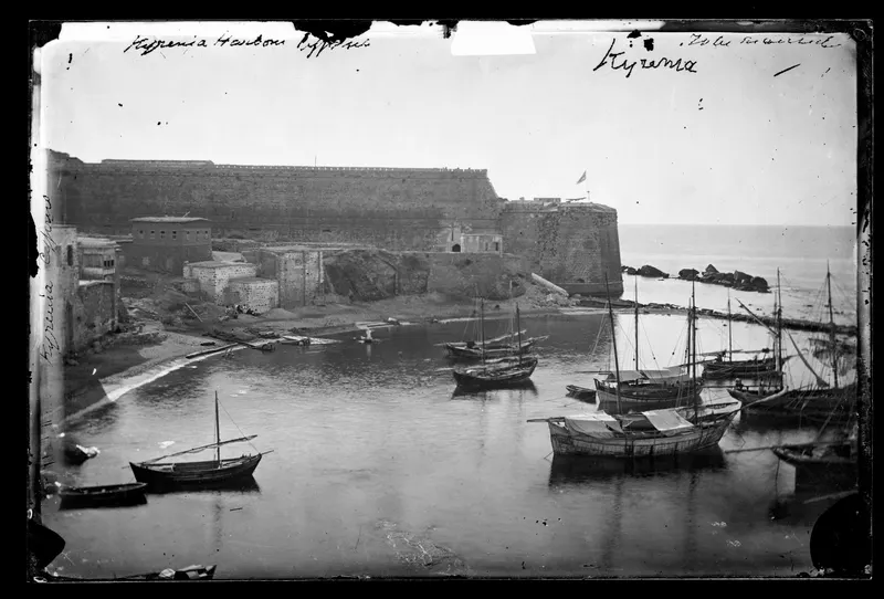 Kyrenia harbour with moored sailing boats in the foreground and a long fortress wall and bastion behind.