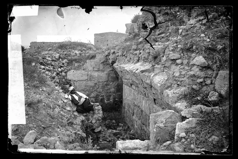 Seated figure in a dark dress beside a trench cut against a massive stone wall, with rubble slopes above