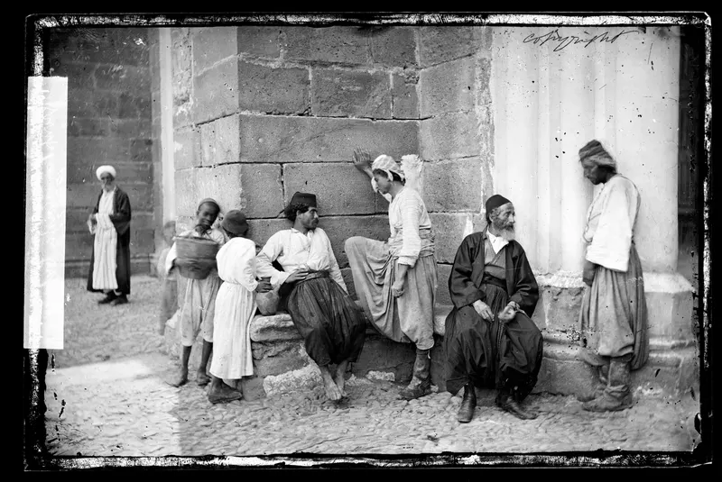 Seven men seated and standing against a stone wall and a fluted column in a cobbled street.