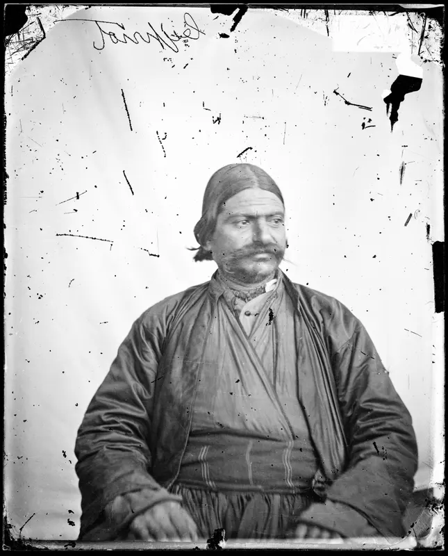 Half-length portrait of a seated man with a large moustache, wearing a long dark coat over a striped sash, against a plain background.