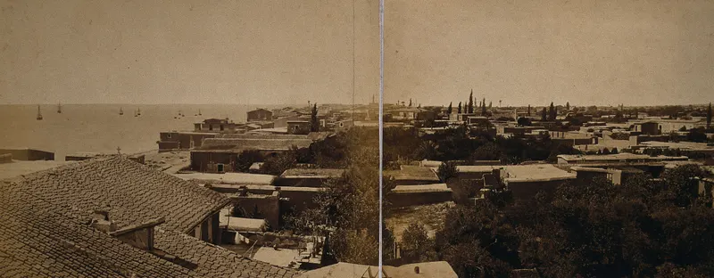 Flat and tiled rooftops of Limasol running down to the sea, with small boats offshore and trees among the houses.