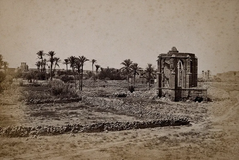 Ruined stone building with a tall pointed window rising above low rubble walls, with palm trees behind it