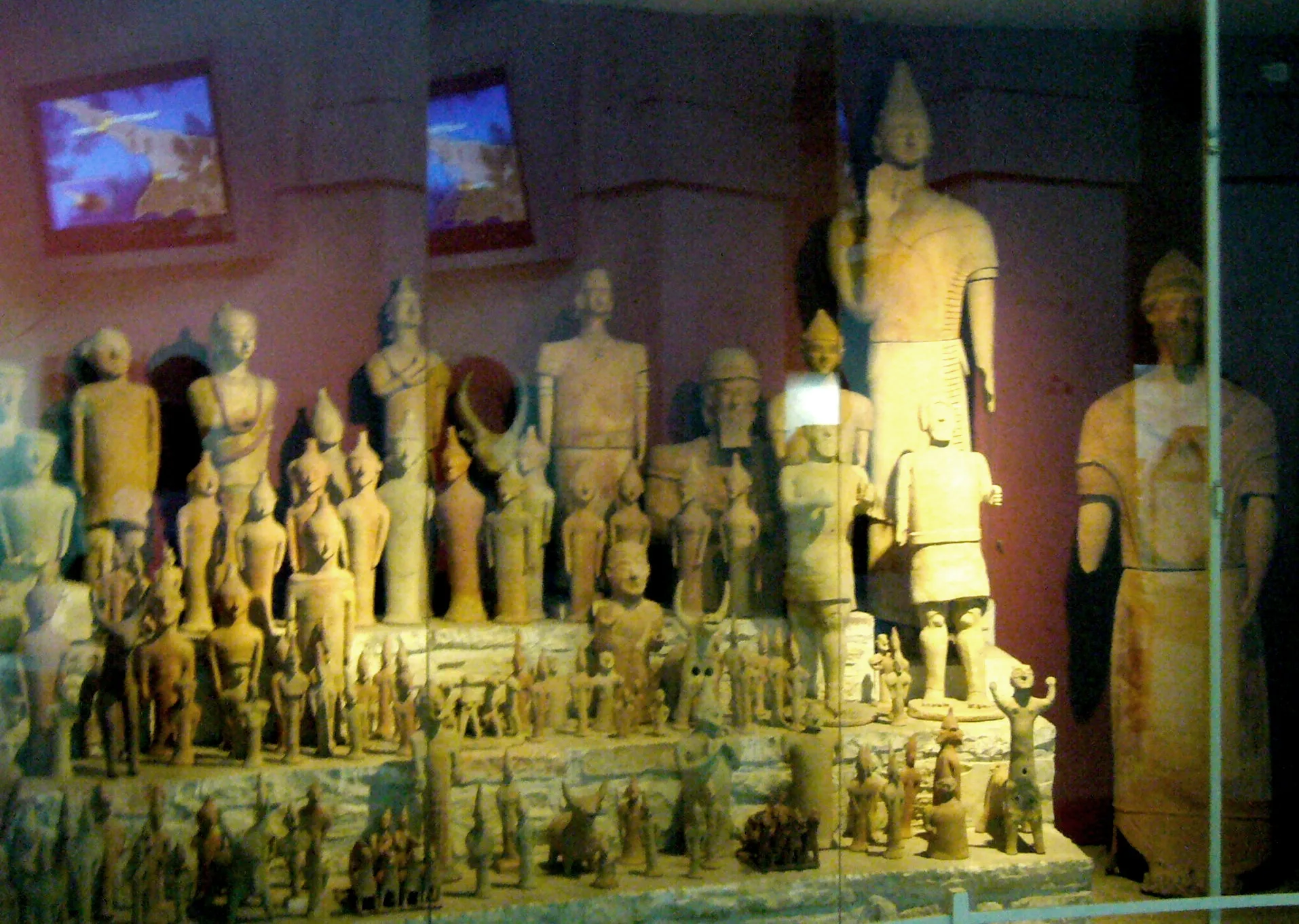 Tiered museum display of terracotta figures from the Ayia Irini sanctuary, small figures massed at the front and life-sized warriors and votaries rising behind a tall helmeted figure at the centre; Cyprus Museum, Nicosia.