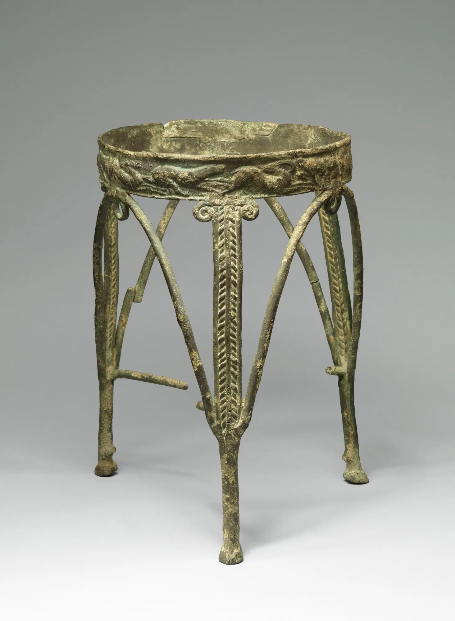 A corroded green bronze tripod stand on three tall splayed legs, its round rim worked with a relief frieze and its central struts with herringbone bands, against a plain gray ground.