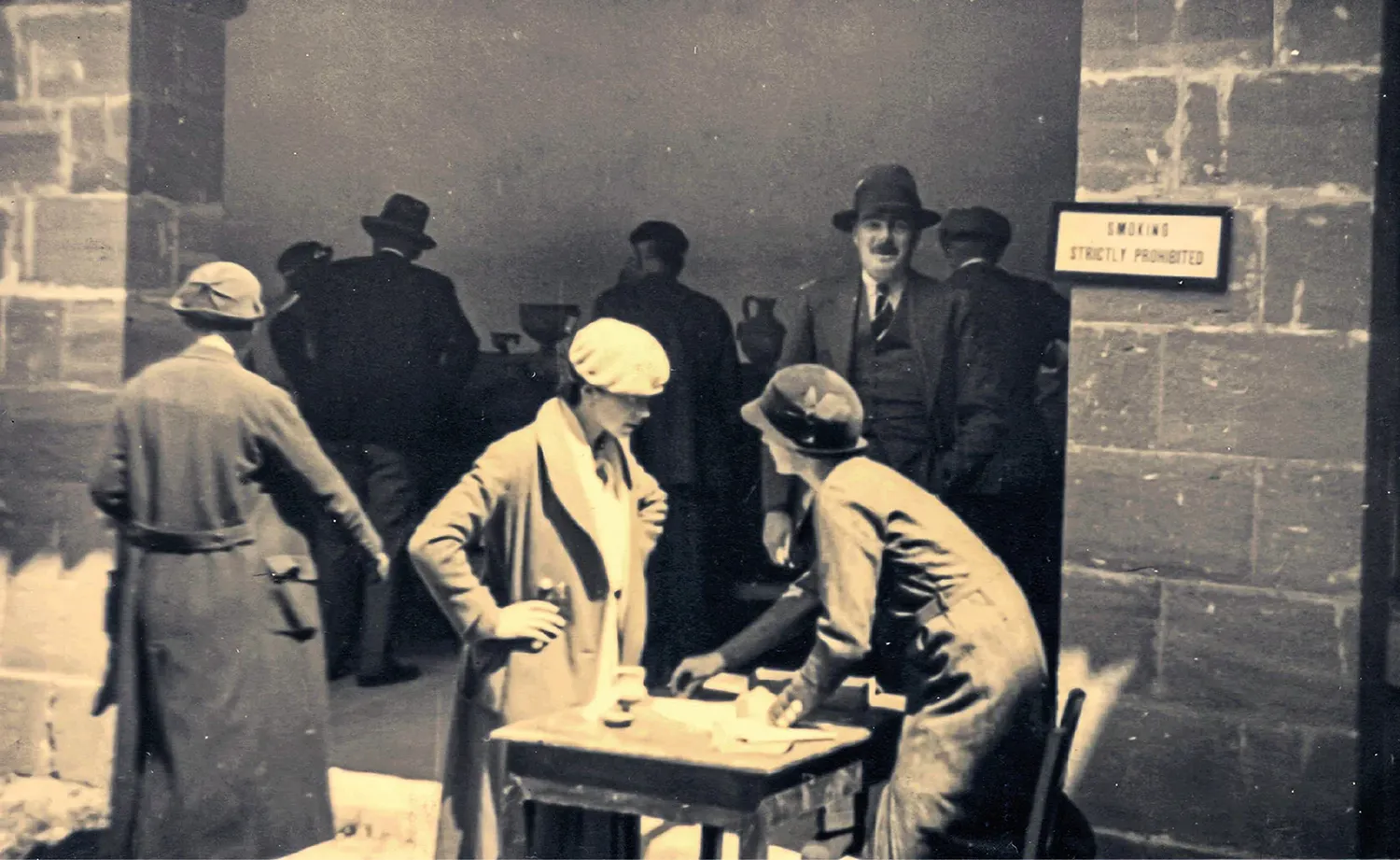 Rupert Gunnis standing behind Joan du Plat Taylor (right) at the Museum saleroom about 1930; the notice on the stone wall reads 'Smoking strictly prohibited.' (Collection of R. S. Merrillees, gift of J. du Plat Taylor; reproduced in Stanley-Price 2025.)