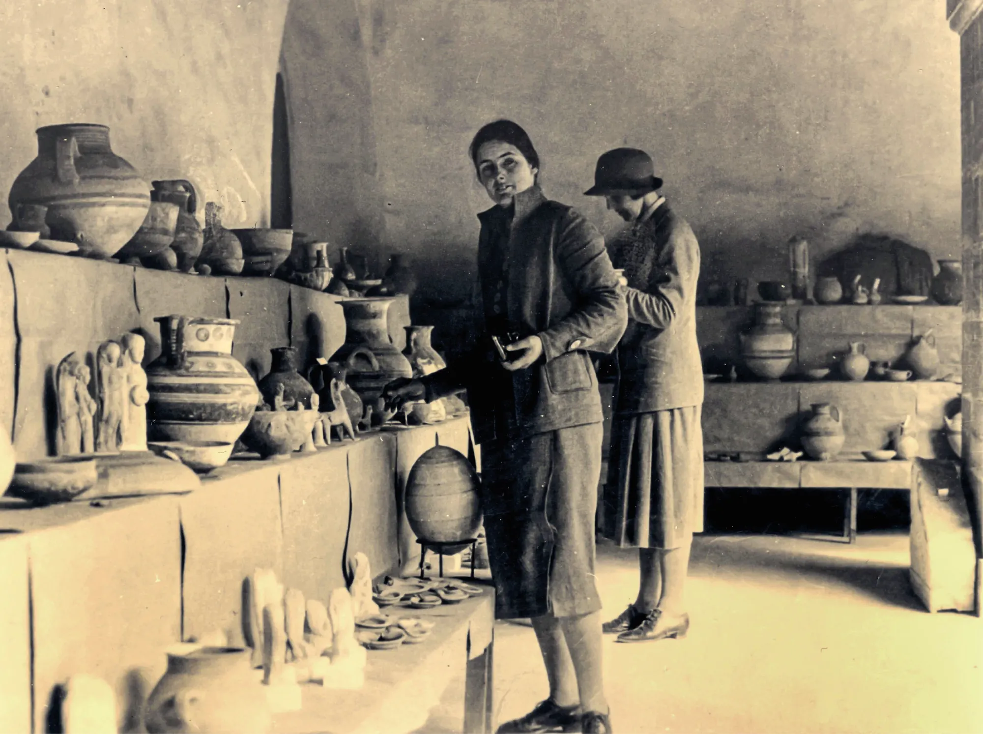 A young woman in a dark coat stands at a long table inside the Cyprus Museum, a small dark vessel in her hands; the table and the shelves behind her are crowded with ancient pots and figurines, and a second woman in a cloche hat stands at her side. About 1930.