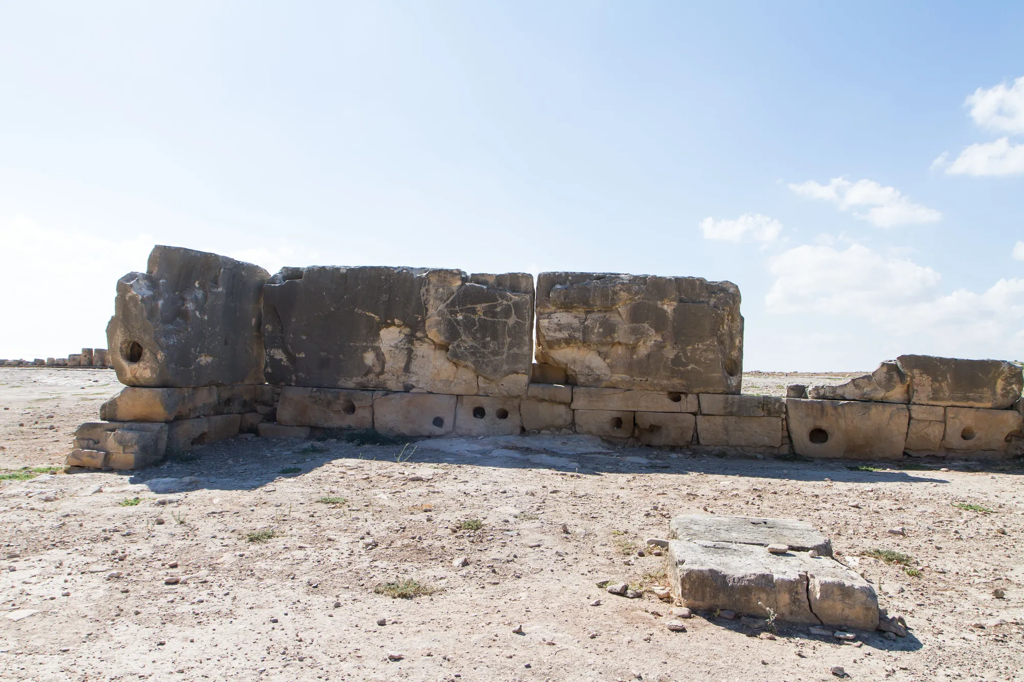 A run of large weathered ashlar blocks pierced by round beam-holes, part of the megalithic sanctuary at Palaepaphos, standing on bare ground under a pale sky.
