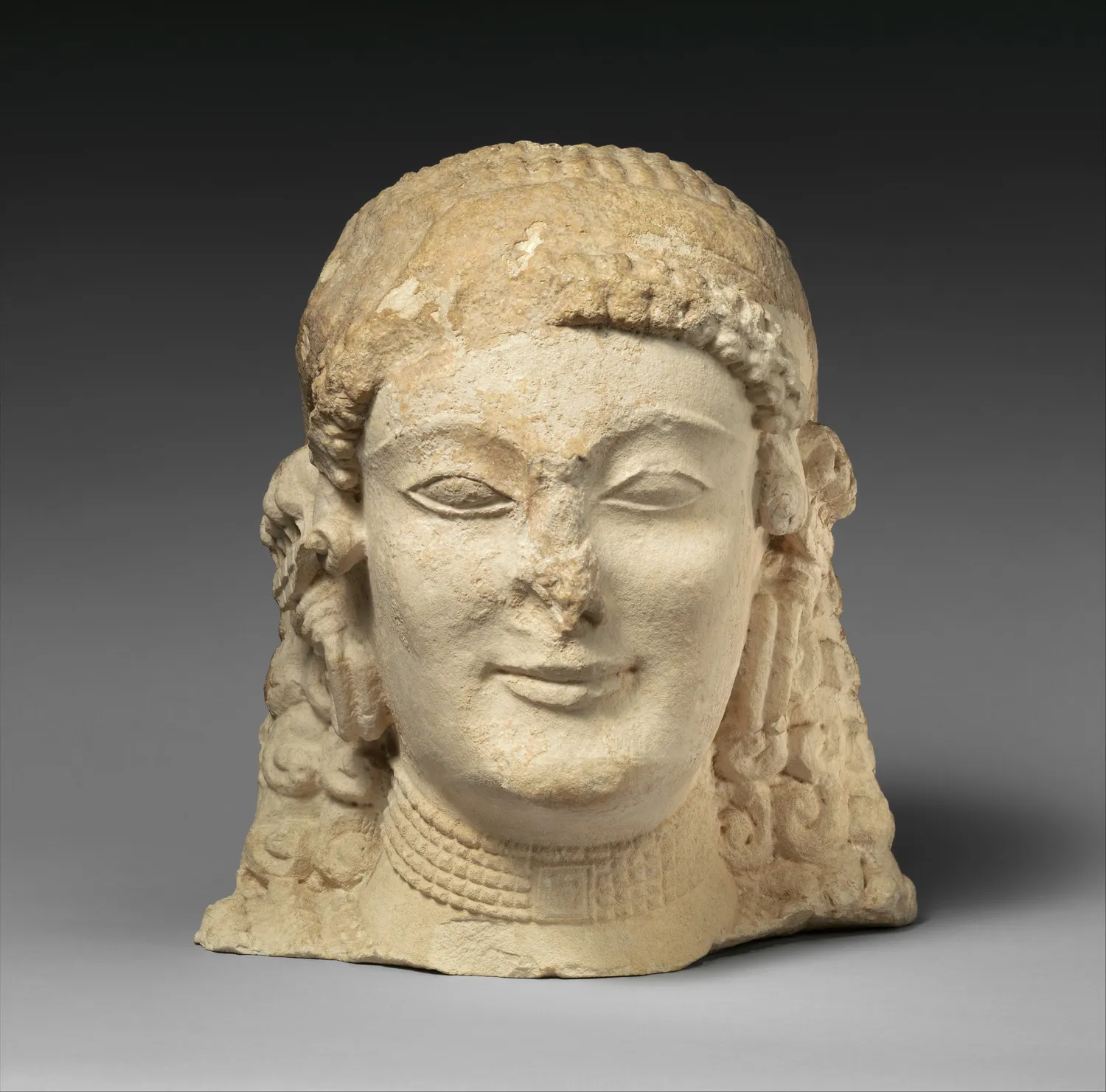 Head of an Archaic Cypriot limestone votary, the hair in rows of curls under a beaded diadem, with a necklace at the broken neck. Metropolitan Museum of Art.