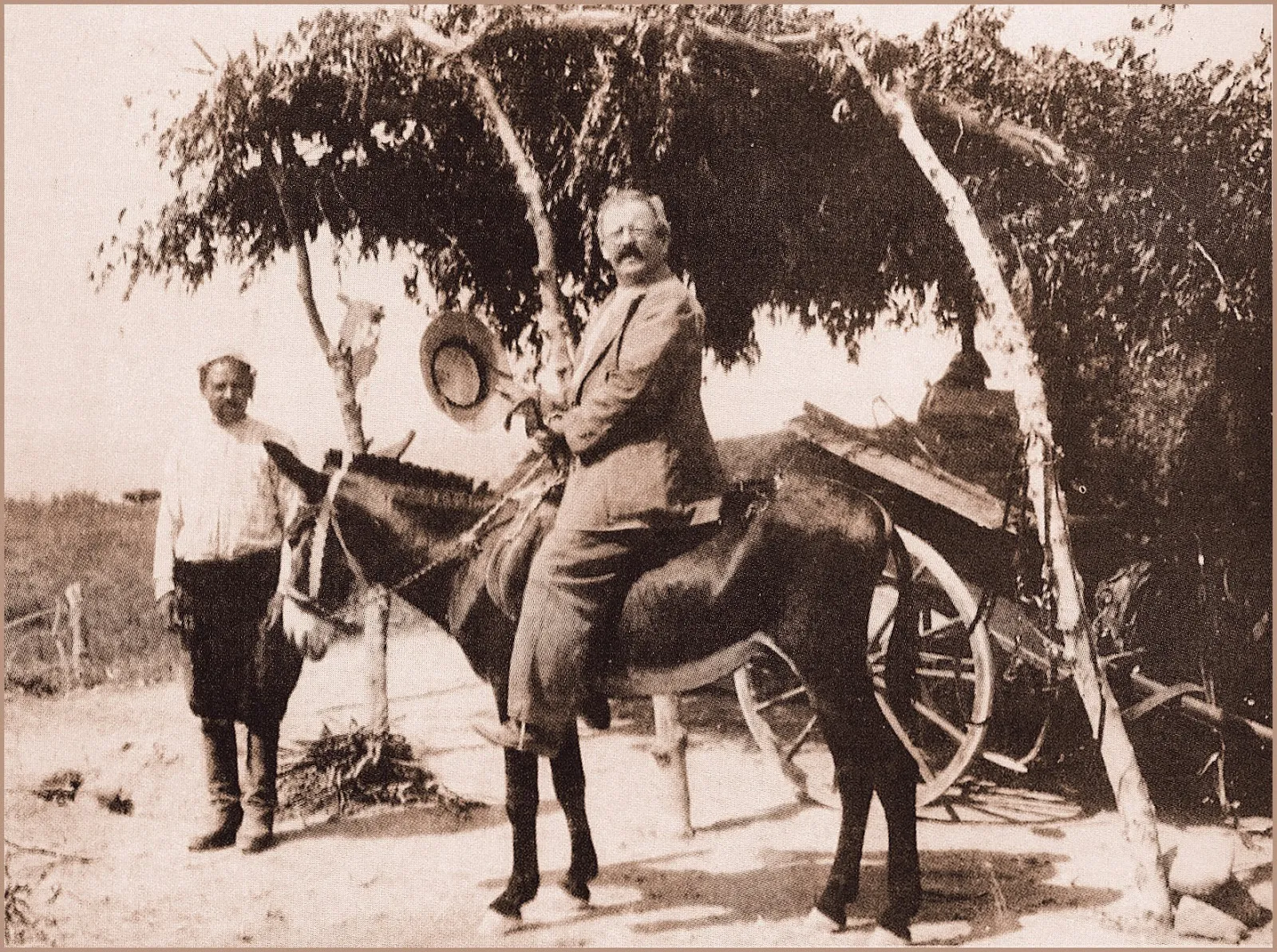 Max Ohnefalsch-Richter, bearded and in a pale field suit, sits astride a mule in the Cypriot countryside in 1910, a local man standing to the left beside a tree and a two-wheeled cart.