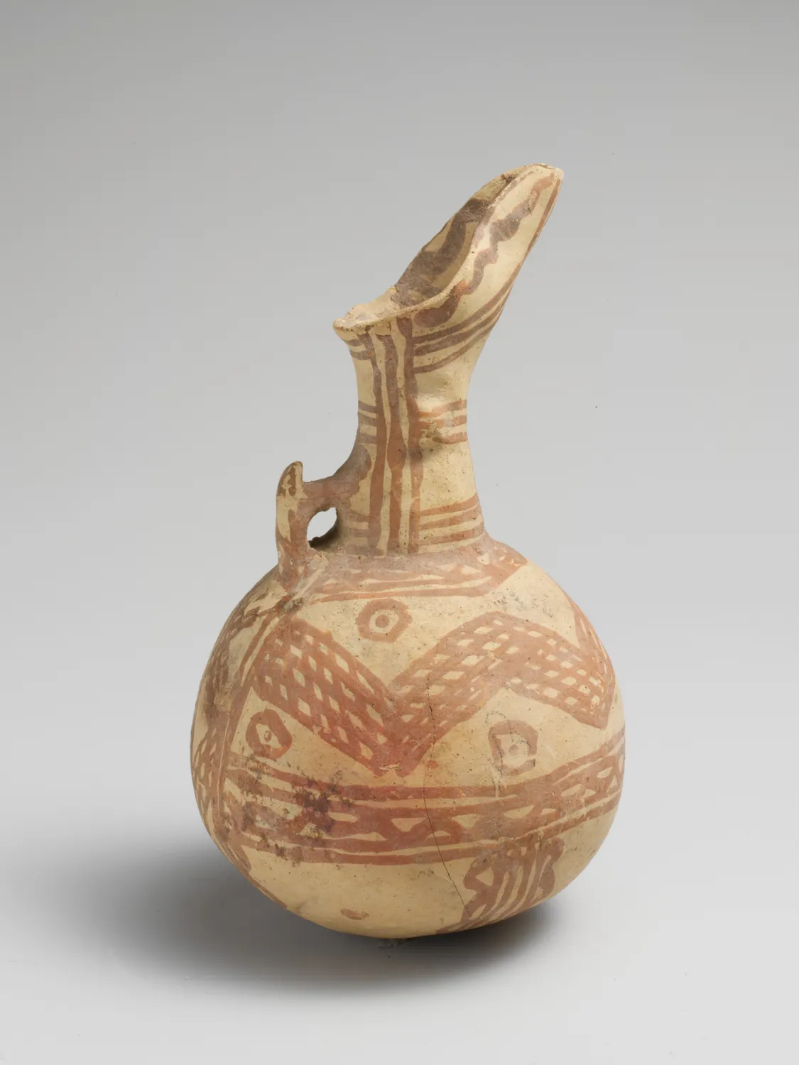 A Cypriot White Painted ware juglet of the Middle Bronze Age, its buff body painted with reddish-brown lattice, zigzag and concentric-circle bands beneath a tall beaked spout. Metropolitan Museum, New York.