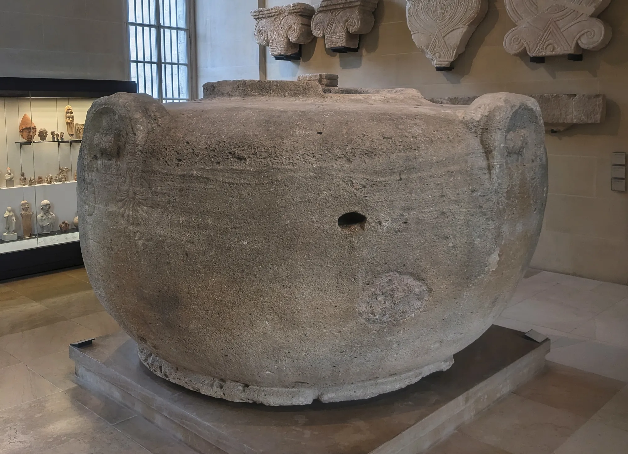 A colossal ancient Cypriot limestone vase with four bull's-head handles, set on a low plinth in a gallery of the Louvre.