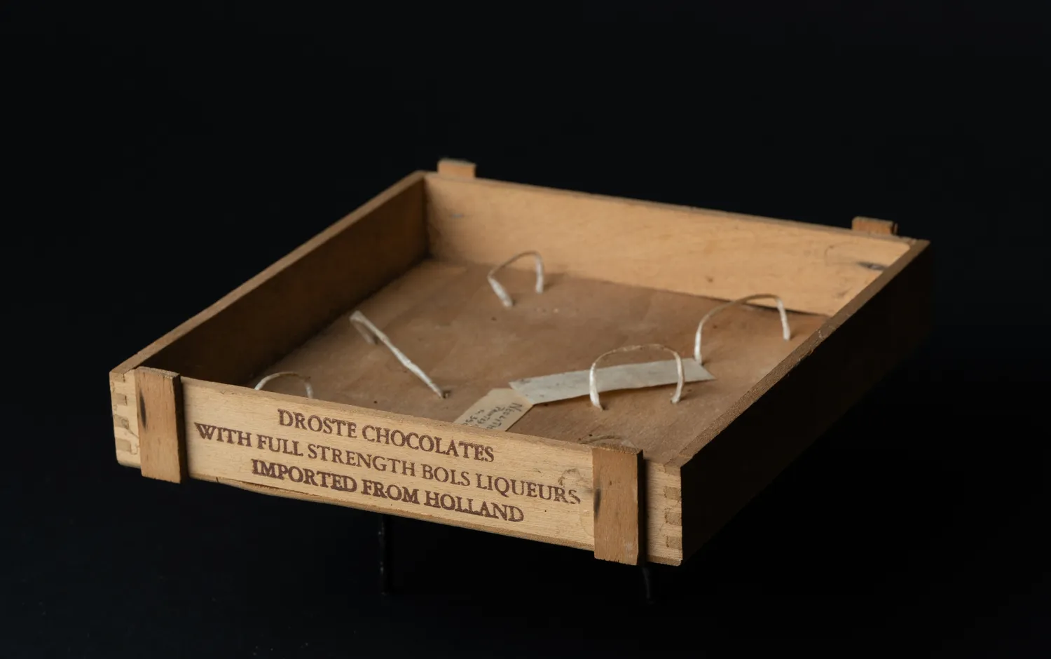The wooden Droste box seen from a low three-quarter angle against black, the branded lettering catching the light along its side, the lid open.