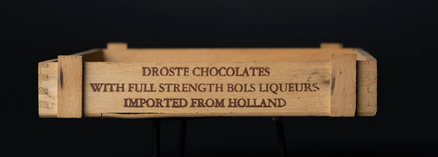 A small pale wooden box photographed end-on against black, its long side branded in faded brown lettering: DROSTE CHOCOLATES, WITH FULL STRENGTH BOLS LIQUEURS, IMPORTED FROM HOLLAND. The corners are finger-jointed.
