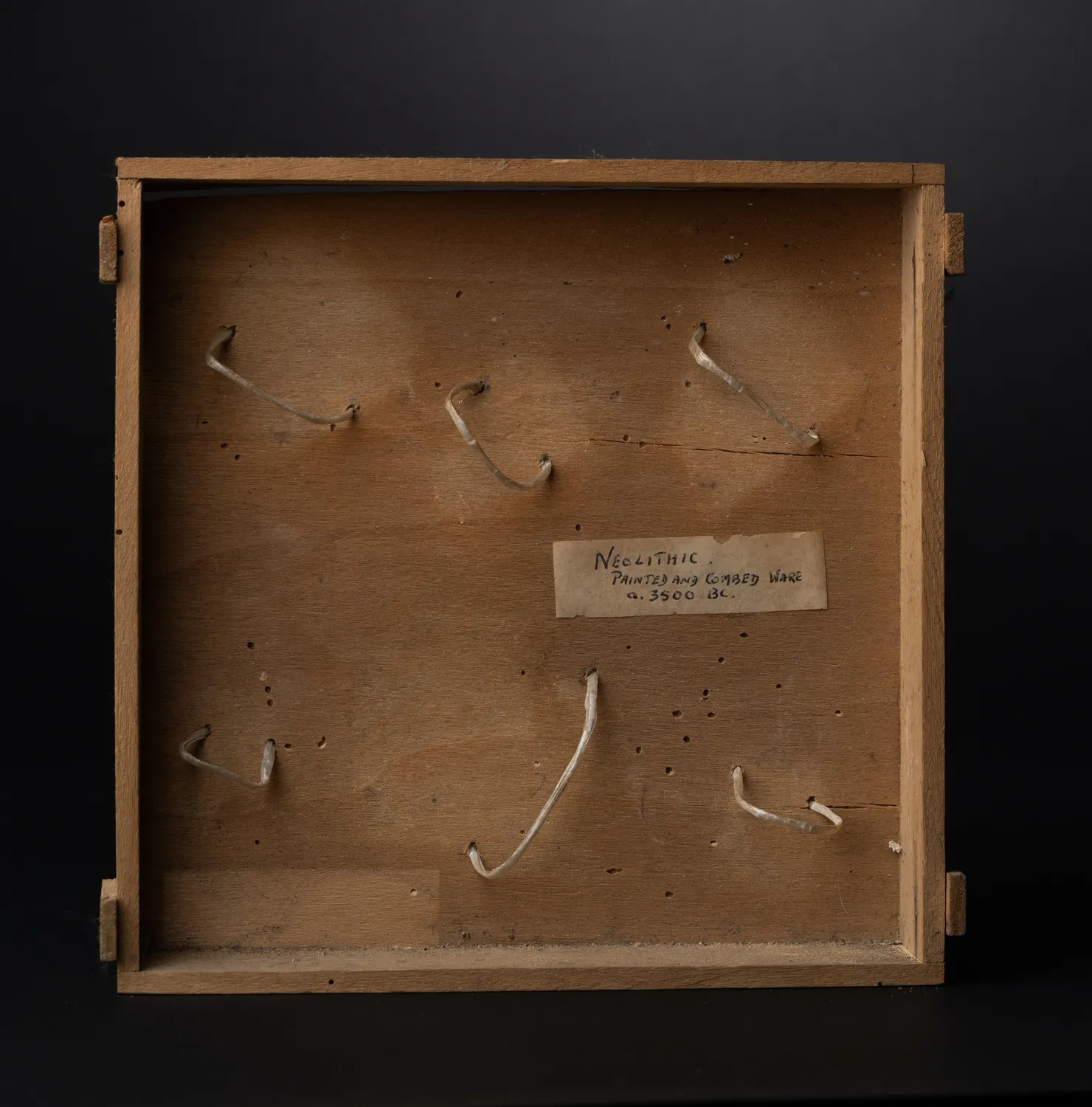 The inside of the box: a thin wooden mount drilled for six bent-wire clips, with the collector's handwritten paper label reading 'Neolithic. Painted and combed ware. c. 3500 BC.' The five sherds have been lifted out of their clips here for photography, and one clip was always left empty.