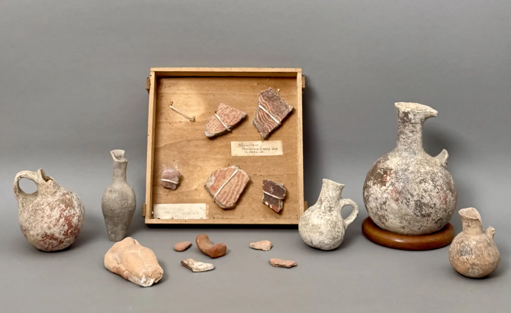 A spread of the judge's Cypriot antiquities laid out on a grey surface: round-bodied Bronze Age jugs and flasks, small juglets, a spouted vessel on a turned wooden stand, and terracotta fragments, arranged around the shallow wooden chocolate box that holds the five clipped Neolithic sherds and its handwritten label.