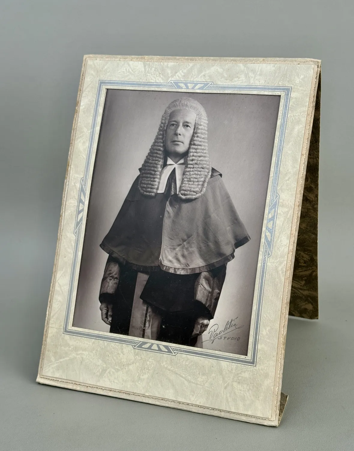 A studio portrait of Judge Griffith-Williams in full judicial dress: a long full-bottomed wig, white lace bands at the throat, a dark silk court coat with a short cape, and knee breeches, mounted on a decorated card easel-frame signed Pavlitis Studio.