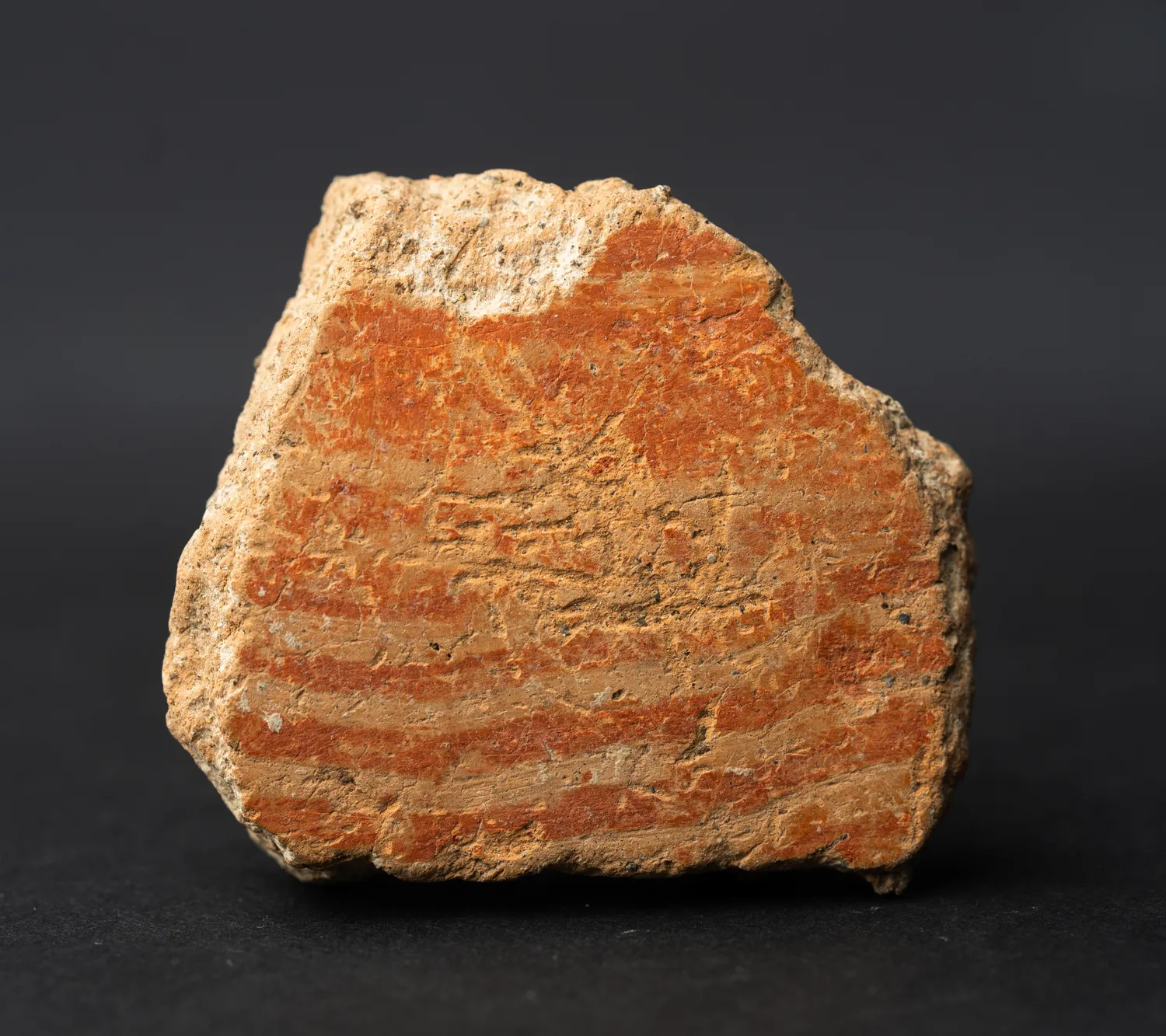 A small reddish potsherd seen from its outer face, the red slip flaking and lime-crusted, with the worn ends of combed bands just visible across the break.