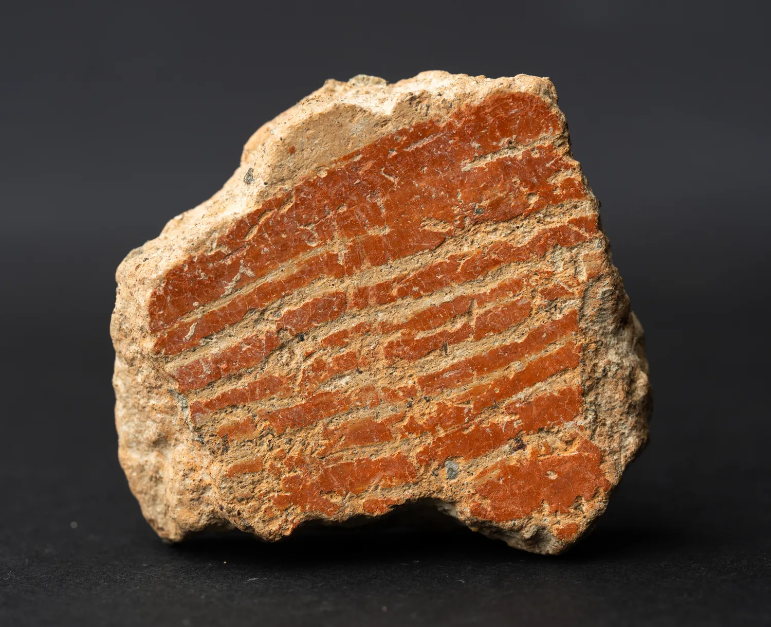 A reddish-brown potsherd with a thick fabric edge, its interior face carrying broad combed bands and heavy whitish lime deposits, photographed against a dark background.