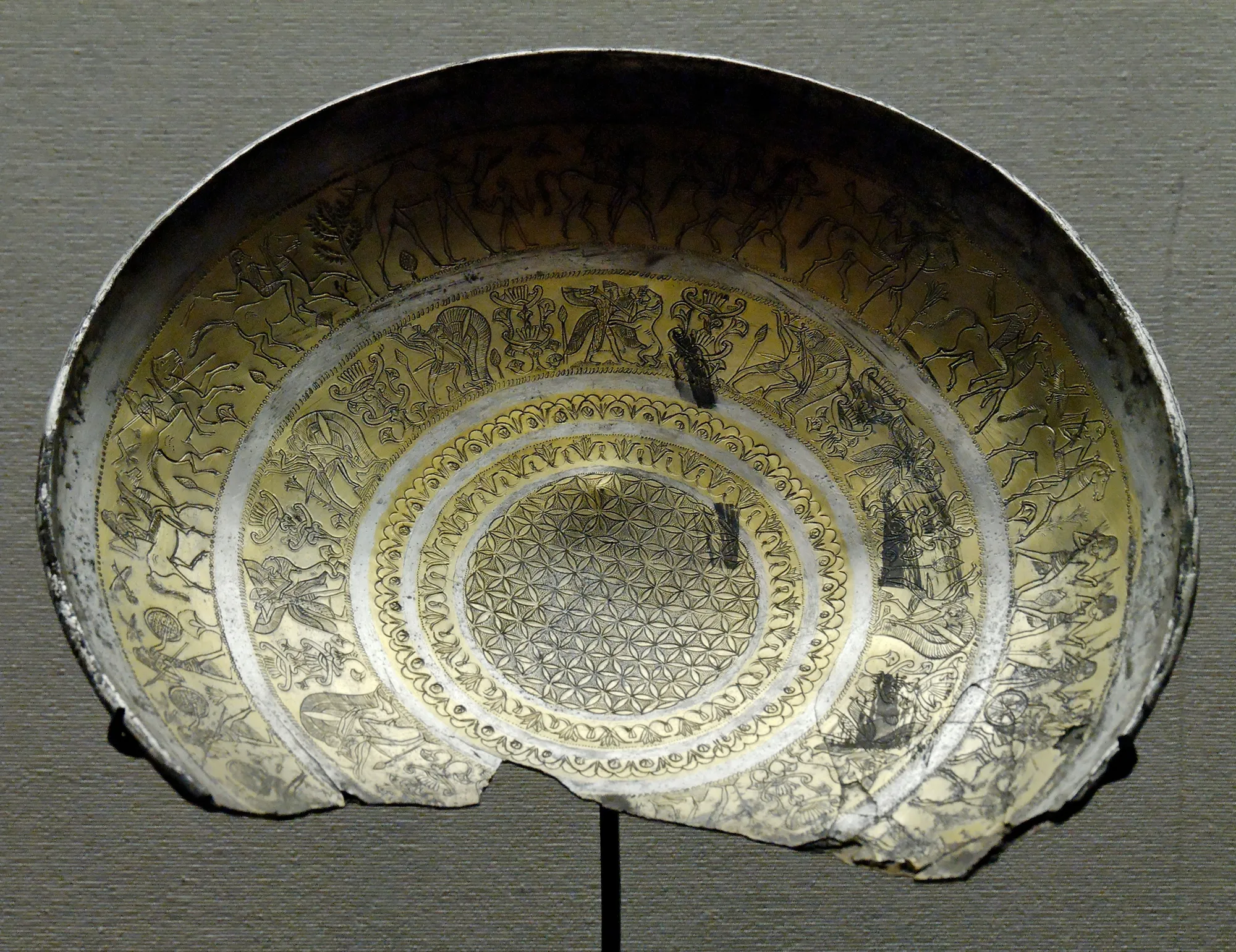 A silver-gilt bowl from Idalion in the Louvre, its interior engraved with concentric friezes of figures, horses and chariots around a central rosette, with a large section of the rim broken away.