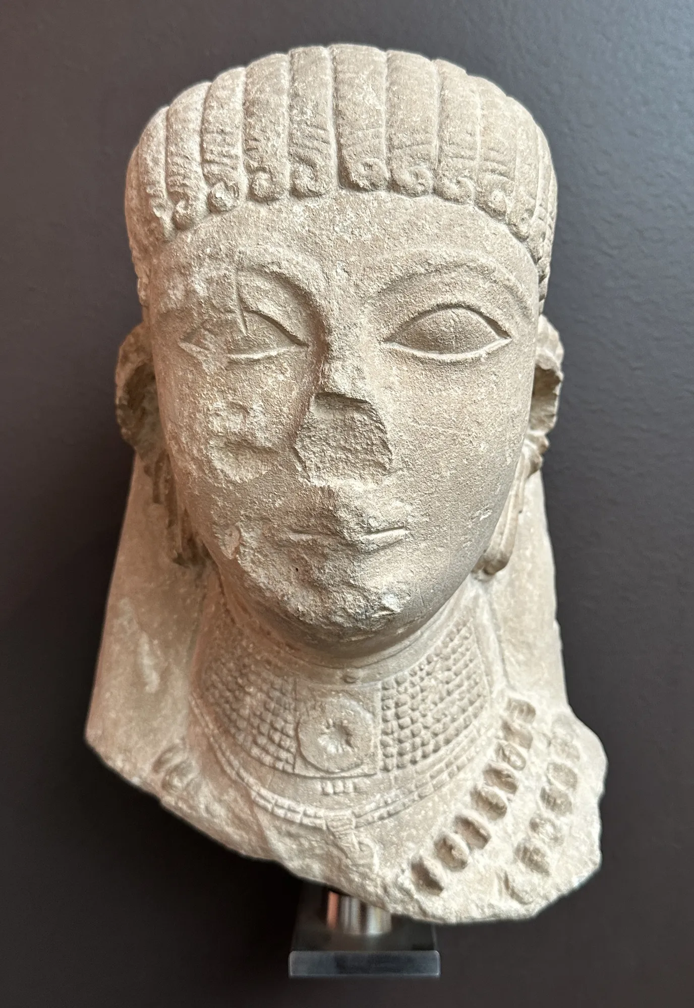 A pale limestone votive head with horizontally banded, beaded hair, large almond eyes and a wide beaded necklace, lit against a dark grey background.