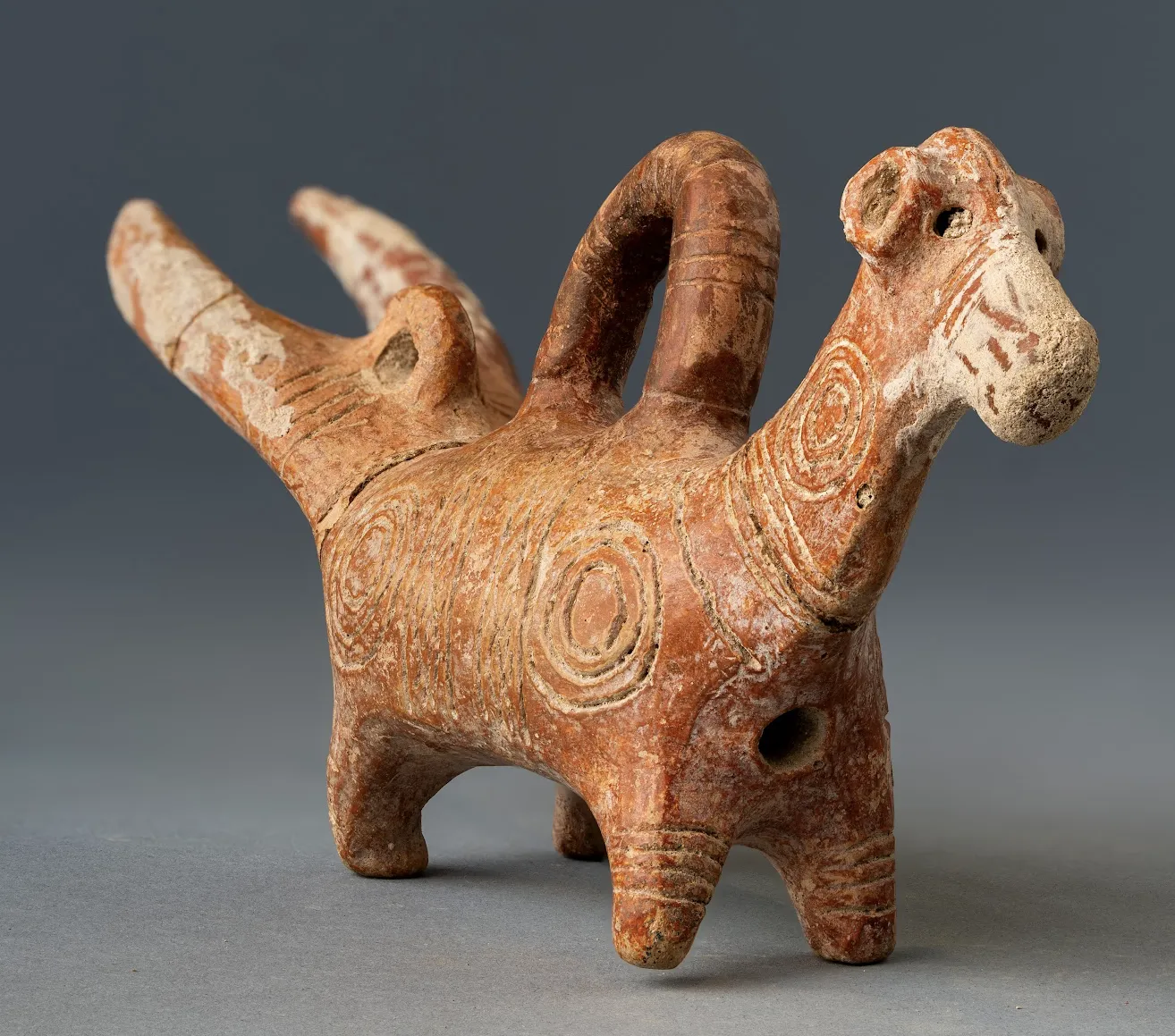 A small Red Polished clay vessel shaped as a four-legged animal, with a white-faced bull's head at one end, a long-eared head at the other, a loop handle over the back, and concentric-circle patterns picked out in white on a red burnished body.