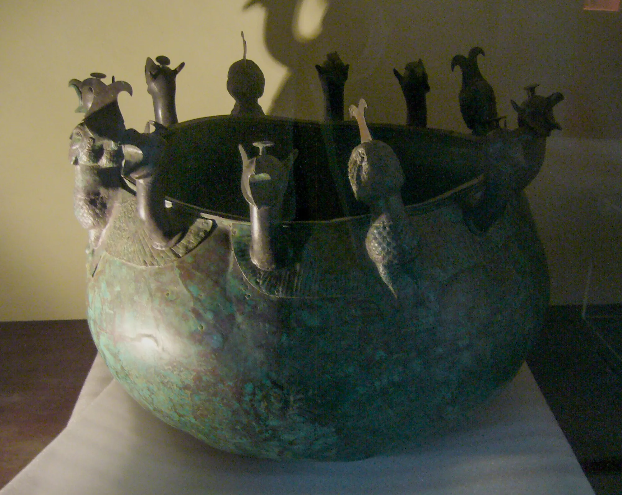 A green-patinated bronze cauldron ringed at the rim with cast griffin heads and winged siren figures, raised on an iron stand in a museum gallery.