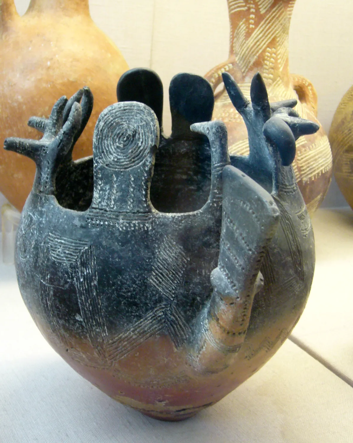 An Early Bronze Age Red Polished vessel with two human figures modelled at its mouth, arms raised over the rim, the body covered in white-filled incised geometric bands. Cyprus Museum, Nicosia (photo Molly, CC BY-SA 4.0).