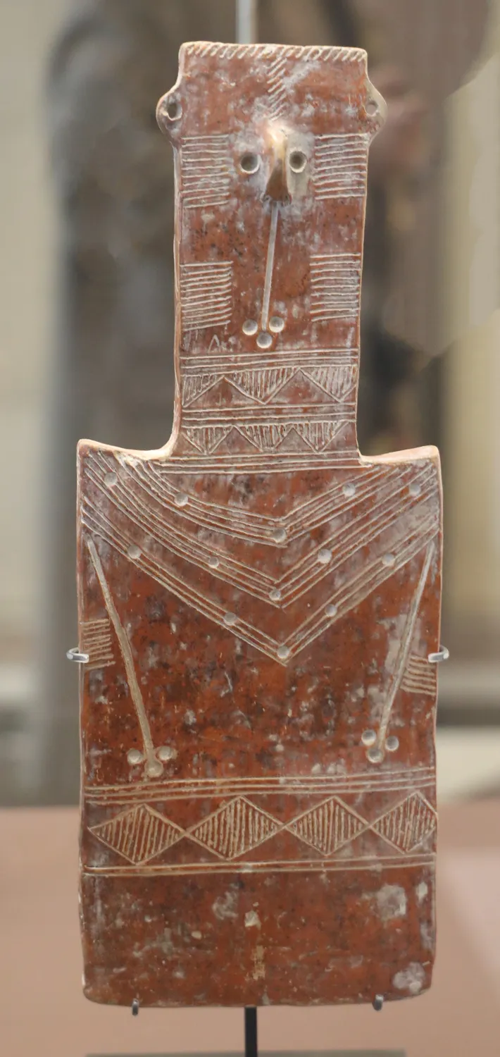 A flat Red Polished plank figure from Vounous: a rectangular plaque with a small head, concentric-circle eyes, pierced ears and a chevron collar picked out in white-filled incision. Louvre, AM 822 (photo Francesco Bini, CC BY-SA 4.0).