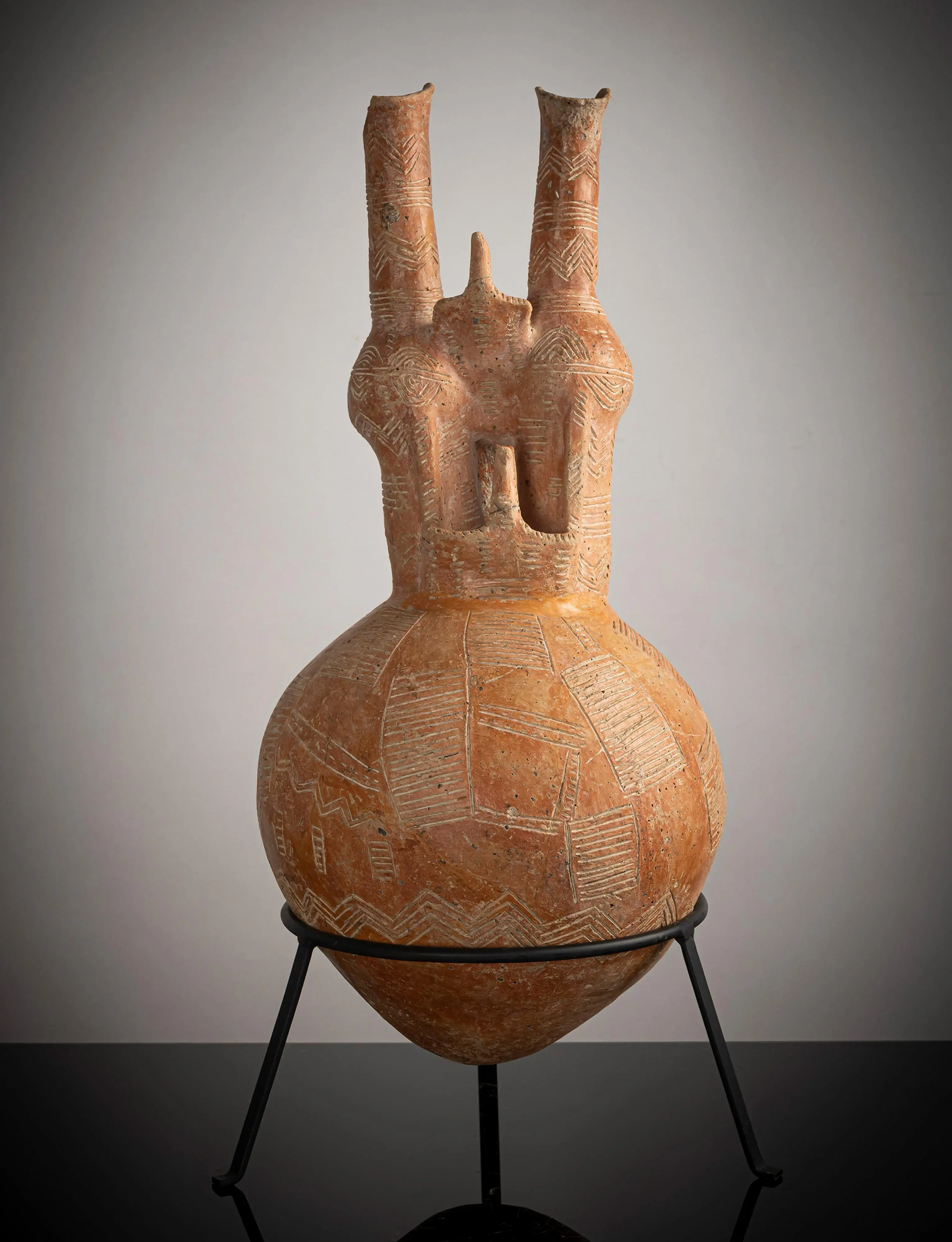 A tall Red Polished Cypriot jug with a near-spherical body covered in white-filled incised geometric pattern, two tall necks rising from the shoulder and two flat, plank-like birds projecting forward, shown on a black metal stand against a grey ground.