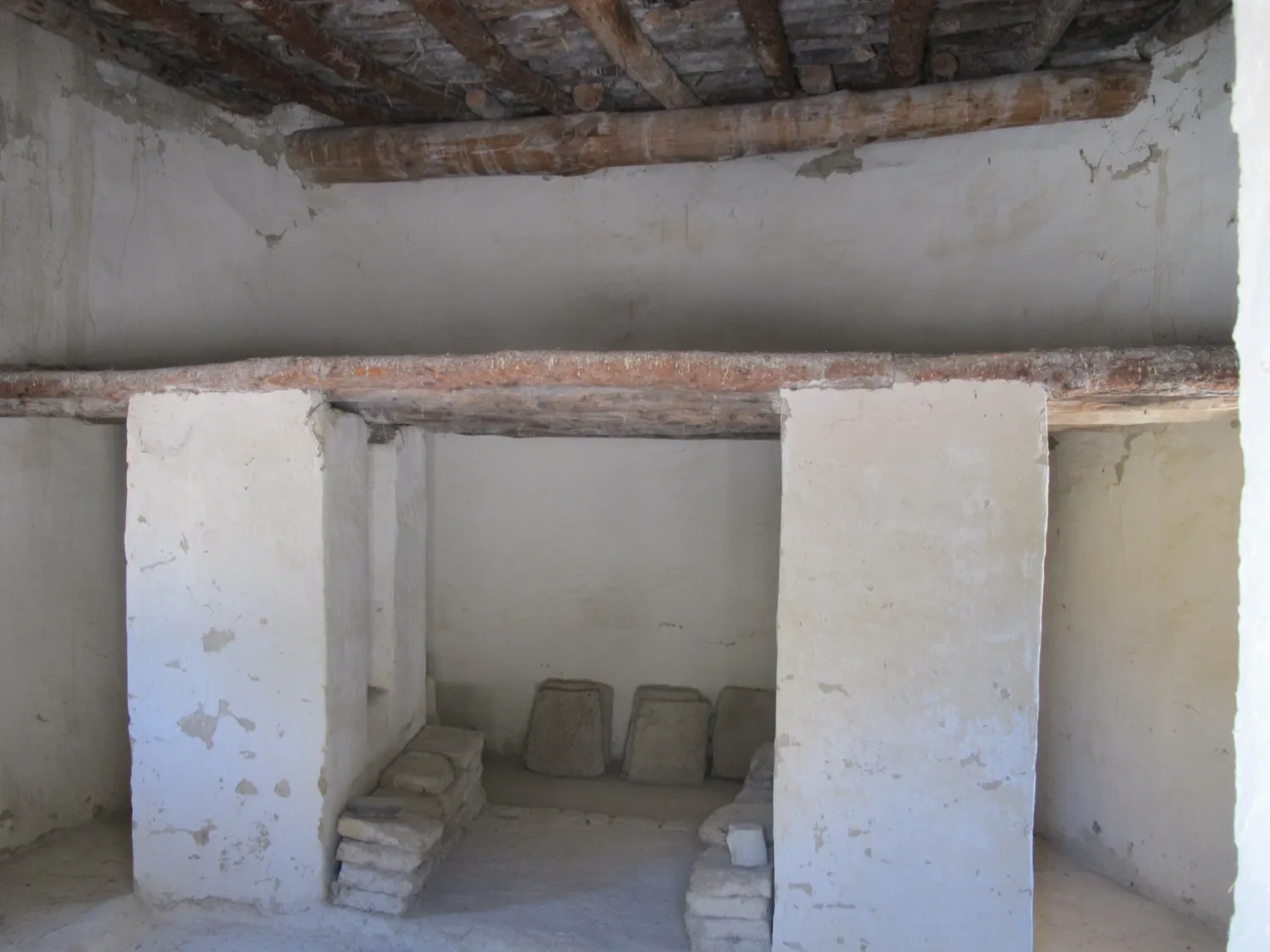Inside one of the replica houses built at Khirokitia in the 1990s: two plastered piers carry a timber beam, and a ceiling of poles closes the room under a flat earth roof. It is a modern reconstruction: the pole ceiling under earth is the kind of roof the burnt S.85 fragments point to, while the piers under the beam are the builders' choice, since Le Brun read such piers as supports for a loft (Lesson 2). Photo: Kritzolina, CC BY-SA 3.0.