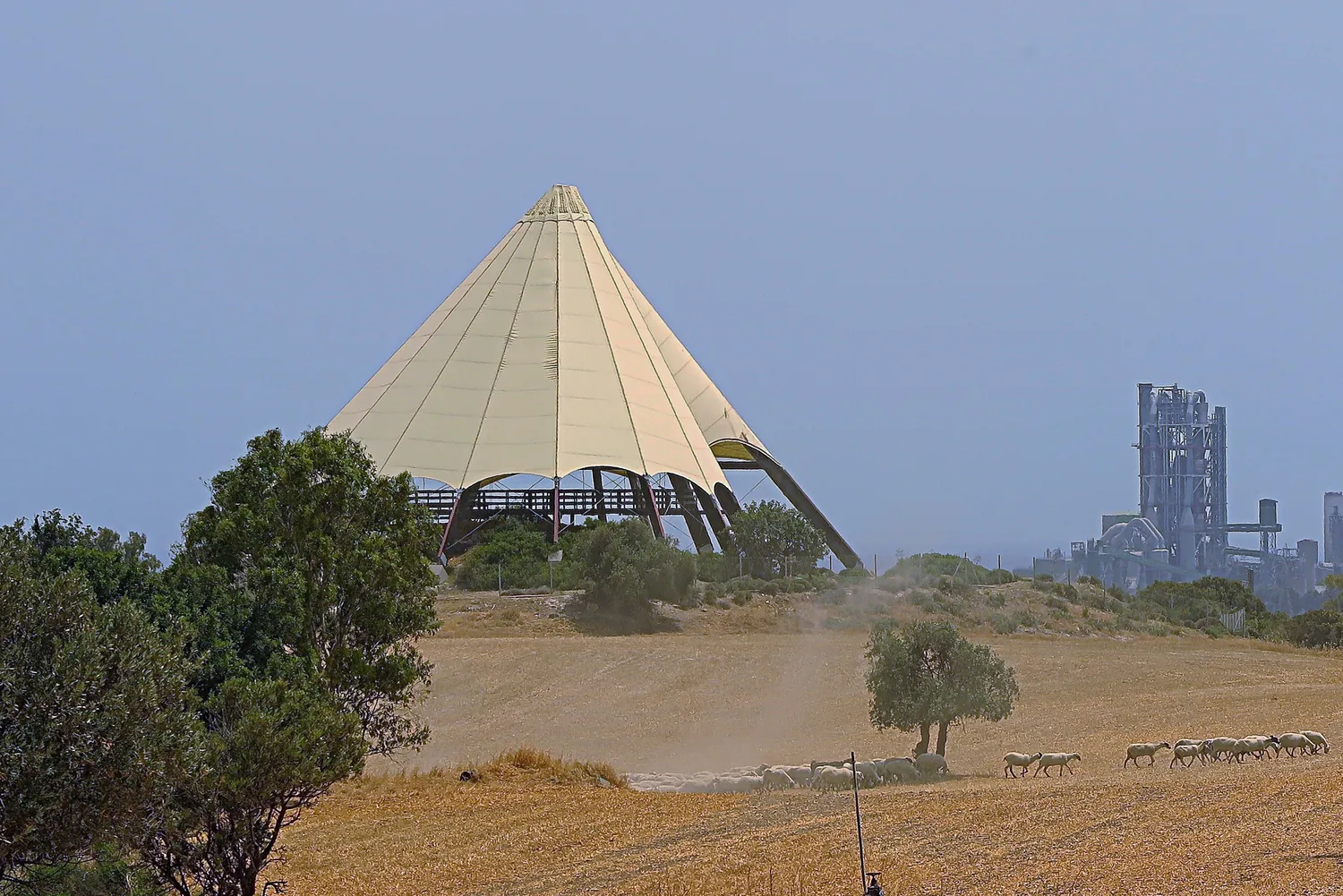 The modern conical shelter over the summit of Kalavasos-Tenta, seen across stubble fields with grazing sheep and a cement works behind. Photo: Rolf Dietrich Brecher, CC BY 2.0.